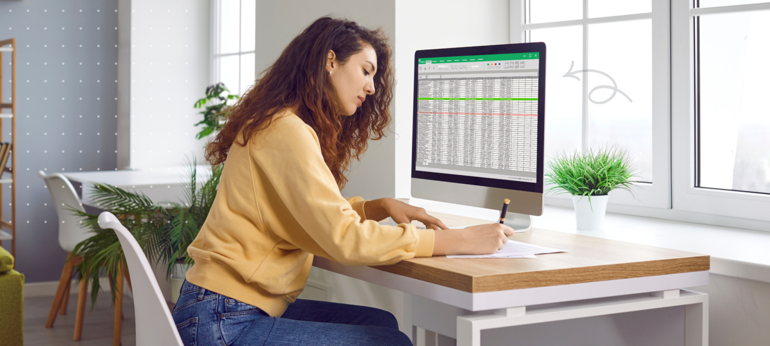 Image of woman sitting at desk, writing on paper in front of a monitor with a speadsheet on the screen
